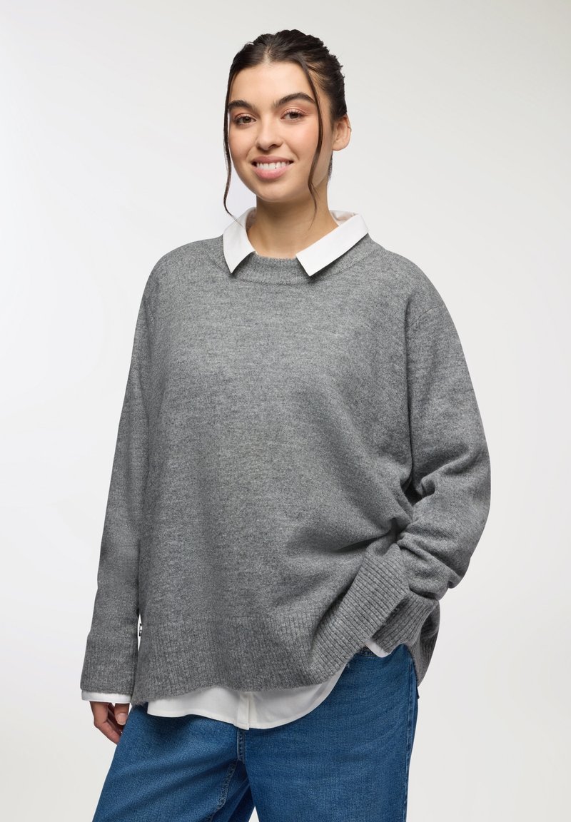 Maglione grigio con una texture morbida, polsini a coste e una vestibilità rilassata, indossato sopra una camicia bianca con colletto, abbinato a jeans in denim blu.