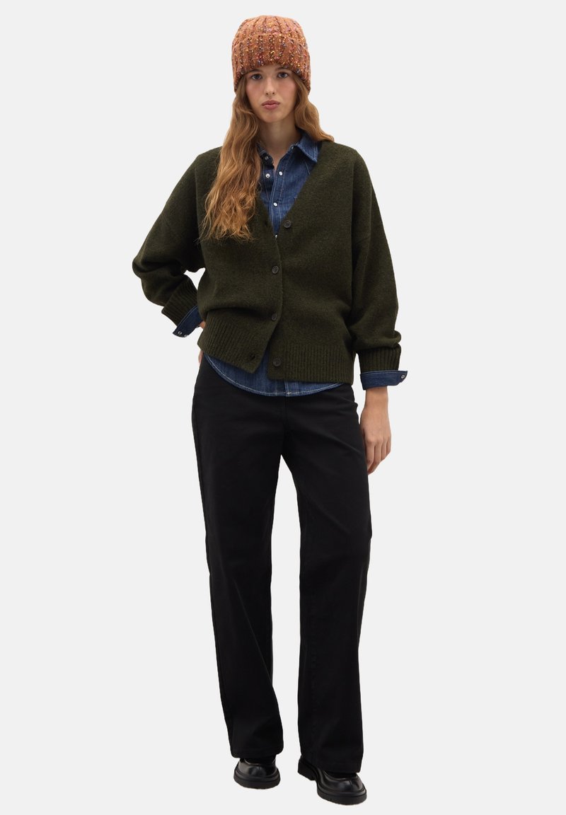 Donna che indossa un berretto in maglia color ruggine, cardigan verde scuro con bottoni, camicia di jeans, pantaloni neri e scarpe nere, in piedi con una mano sulla vita.