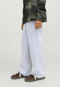 Jeans de pierna ancha en azul claro, con textura de denim, que presentan un ajuste relajado. Combinados con zapatillas marrones que tienen rayas laterales blancas.