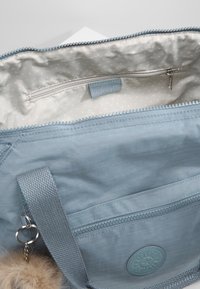 Sac en tissu bleu clair avec fermeture éclair, doublure intérieure à pois, poche zippée, patch logo et porte-clés pompon en fourrure attaché à la poignée.