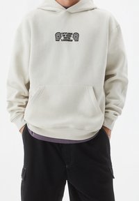 Sudadera beige claro con un bolsillo frontal y puños acanalados. Presenta un diseño gráfico negro en el pecho. Material de algodón, corte relajado.