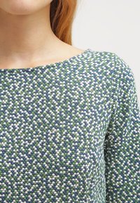 Kuviollinen paita, jossa on vihreitä, sinisiä ja valkoisia pisteitä teksturoidussa kudoksessa; leveä veneen kaula-aukko ja istuvat hihat.