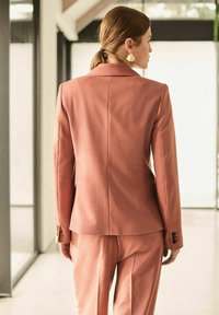 Blazer tailleur rosa pallido con una vestibilità strutturata, colletto a rever, spacco posteriore singolo e dettagli con bottoni sulle maniche, abbinato a pantaloni coordinati.