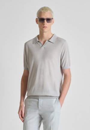 Joven con cabello rubio corto que lleva gafas de sol grises, camisa polo de manga corta gris claro y pantalones grises claros frente a un fondo liso.