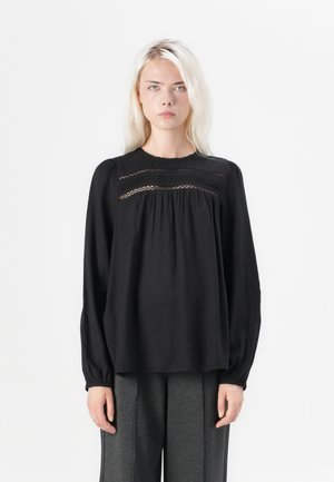 Schwarze Bluse mit langen Ärmeln und rundem Ausschnitt, mit Spitze entlang der Vorderseite und gerafftem Stoff, kombiniert mit grauen Hosen.