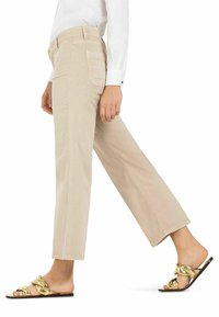 Beige Baumwollhosen mit geradem Bein, ausgestattet mit Gesäßtaschen und Knopfverschluss. Kombiniert mit goldenen Rutsch-Sandalen.