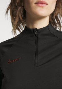Schwarzes sportliches Zip-Up-Oberteil mit hohem Kragen, mit einem kleinen orangefarbenen Nike-Swoosh-Logo auf der linken Seite. Glattes, strukturiertes Material.