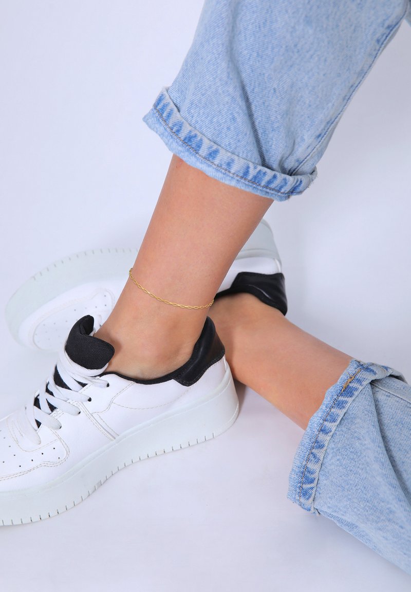 Hvide sneakers med sorte detaljer, rund tå, tykk sål, parret med oprollede lyseblå denim jeans og en tynd guld ankelkæde.