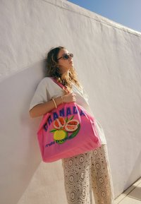 Borsa tote rosa con grafica di melograno e limone, etichettata "GRANADA Fruta Tropical." Modella indossa abbigliamento chiaro e occhiali da sole rotondi.
