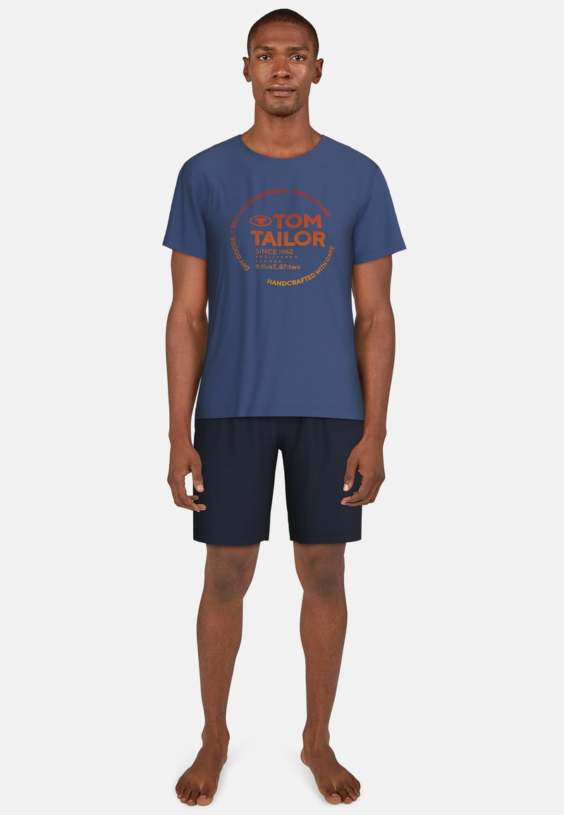 Camiseta de algodón azul marino con gráfico circular en naranja, mostrando el nombre de la marca "TOM TAILOR" y texto. Combinada con pantalones cortos azul marino.
