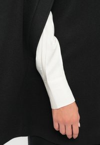 Un capo d'abbigliamento scuro che pende sopra una camicia bianca a maniche lunghe con finitura lucida ai polsini. Tessuto liscio con dettagli di sartoria sottili.
