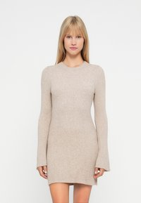 Mujer vistiendo un vestido mini de punto acanalado beige de manga larga, de pie frente a un fondo blanco liso.