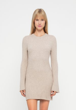 Mujer vistiendo un vestido mini de punto acanalado beige de manga larga, de pie frente a un fondo blanco liso.