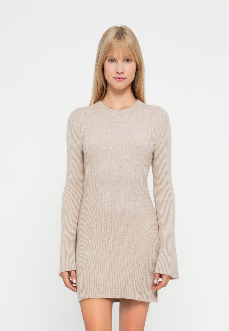Mujer vistiendo un vestido mini de punto acanalado beige de manga larga, de pie frente a un fondo blanco liso.