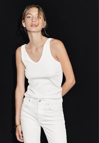 Bílý bavlněný top bez rukávů s výstřihem do V je kombinovaný s bílými džíny. Pozadí je jednobarevné černé, což zvýrazňuje barvy oblečení.