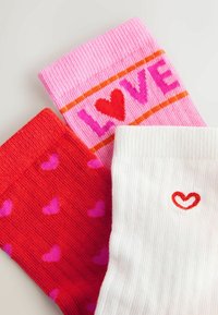 Trīs krāsainas zeķes ar sirsniņu rakstiem: rozā ar tekstu "LOVE", sarkana ar mazām rozā sirsniņām un balta ar nelielu sarkanu sirsniņu.