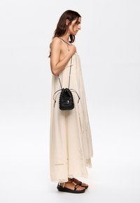 Jasnobeżowa maxi sukienka z haftowanymi detalami, zestawiona z czarną plecioną torebką typu bucket oraz brązowymi sandałami z paskami.