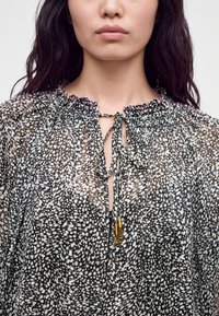 Femme portant un chemisier transparent noir et blanc à pois, avec un col froncé noué par de fines ficelles se terminant par des perles dorées.