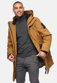 Chaqueta aislante marrón con capucha, cierre de cremallera, forro de forro polar y detalles negros. Combinada con un suéter oscuro y pantalones grises.