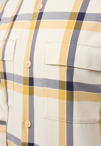 Korte mouwen geruite blouse in crème, met gele, marineblauwe en grijze strepen. Twee borstzakken en knoopsluitingen voor detail. Glad stoftextuur.