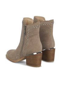Beżowe zamszowe botki za kostkę z bocznym zamkiem błyskawicznym, detalami w postaci ćwieków oraz brązowym obcasem przypominającym drewno.