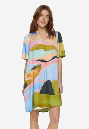 Nainen pukeutunut löysään, lyhythihaiseen mekkoon, jossa on värikäs abstrakti maisemakuosi pastellisävyissä ja maanläheisissä väreissä.