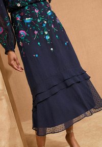 Fustă bleumarin, până la genunchi, cu volane suprapuse, material texturat, broderie florală detaliată în roz și turcoaz, suprapunere transparentă.
