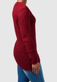 Maglione rosso lavorato a maglia con un motivo testurizzato, maniche lunghe e polsini a coste, abbinato a jeans di denim. La vista laterale mostra il fit e i dettagli del design.