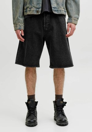 Person iført sorte denimshorts, sorte snørestøvler, mørke sokker og en falmet denimjakke, der står mod en ensfarvet baggrund.