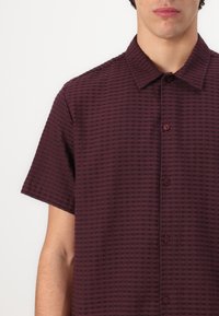 Korte mouwen, bordeauxrood shirt met een gestructureerd ruitpatroon. Voorzien van een puntige kraag en bijpassende knopen langs de voorkant.