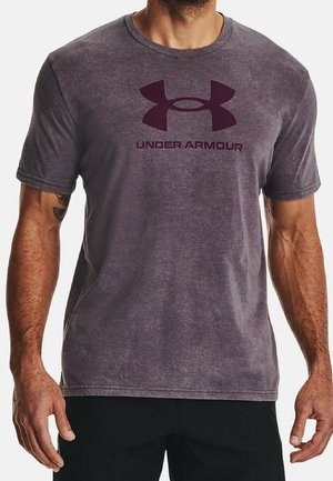 Mann trägt ein ausgewaschenes lila Under Armour T-Shirt und schwarze Hose und steht mit entspannt an den Seiten hängenden Armen.