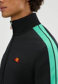 Chaqueta negra de cremallera con un cuello alto. Presenta franjas en color teal que contrastan y un logo circular en el pecho. Textura de tela suave.