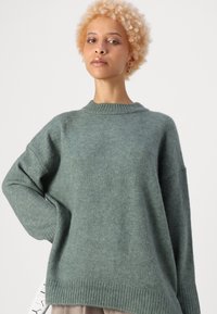 Maglione verde lavorato a maglia con scollatura tonda, spalle abbassate e orlo a costine. Texture morbida, vestibilità rilassata e design leggermente oversize.