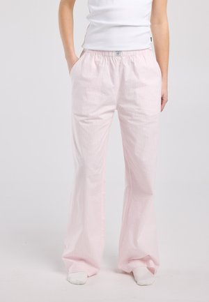 Persoon die losse, lichtroze en witte broek draagt met verticale strepen, een elastische tailleband en zakken, gecombineerd met witte sokken en een wit shirt.