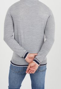 Szary sweter dzianinowy z okrągłym dekoltem, dopasowane mankiety z granatowymi i białymi paskami, w połączeniu z niebieskimi dżinsami, widok z tyłu.