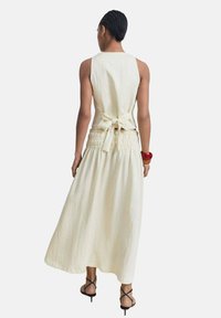 Vestido maxi sin mangas, fluido, en tela color crema con cintura fruncida y detalle de lazo en la parte posterior, combinado con tacones negros con tiras.