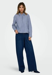 Gestreifte Hemdbluse in Blau und Weiß mit Kragen und einer Brusttasche, kombiniert mit weit geschnittenen dunkelblauen Jeans und spitzen schwarzen Schuhen.