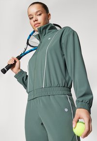Grønn atletisk treningsoverall med hvite detaljer, med høy krage og elastisk midjebånd, holdt av en person med en tenniskølle og ball.
