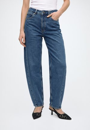 Modèle portant un jean bleu ample à jambes droites, un haut blanc et des escarpins noirs pointus, une main dans une poche.