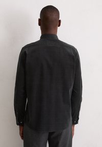 Homme aux cheveux courts portant une chemise noire à manches longues, vu de dos, devant un mur de couleur claire.
