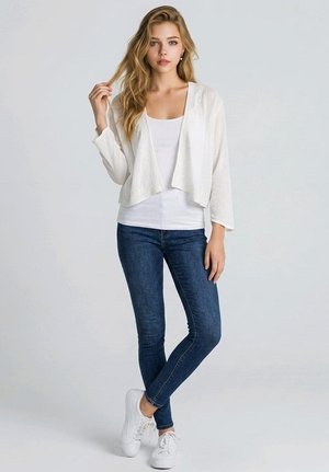 Joven mujer con cabello rubio largo, vistiendo un cárdigan blanco, top blanco, vaqueros ajustados azules y zapatillas blancas, de pie frente a un fondo liso.