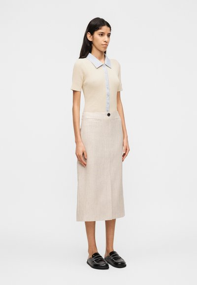 Μια γυναίκα στέκεται στραμμένη ελαφρά προς τα δεξιά, φορώντ ας ένα μπεζ κοντομάνικο τοπ με γιακά, μπεζ midi φούστα με κουμπιά μπροστά και μαύρα παπούτσια χωρίς κορδόνια.