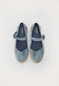 Sapatos de tecido azul com sola plana, uma caixa de dedos larga e uma tira ao longo do peito do pé. Possuem um padrão texturizado e uma sola ao estilo espadrille.