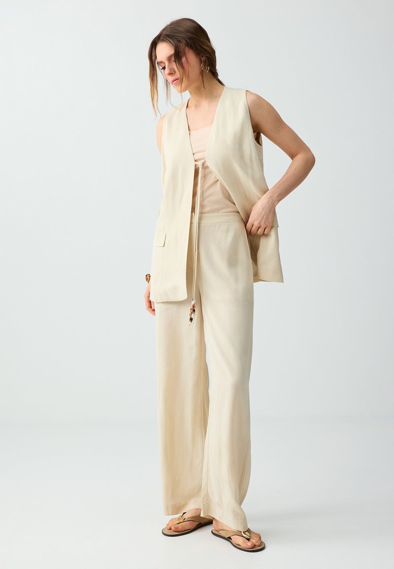 Femme ajustant un gilet sans manches beige, portant un pantalon large assorti et des sandales, debout devant un fond uni clair.