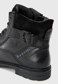Botină din piele neagră până la gleznă, cu partea superioară din suede, detalii cu cataramă metalică și talpă din cauciuc texturată. Logo-ul vizibil pe lateral.