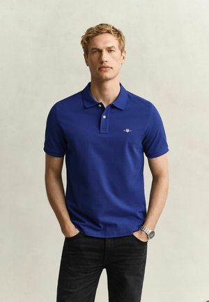 Homme blond debout les mains dans les poches, portant un polo bleu et un pantalon noir, fond clair uni.