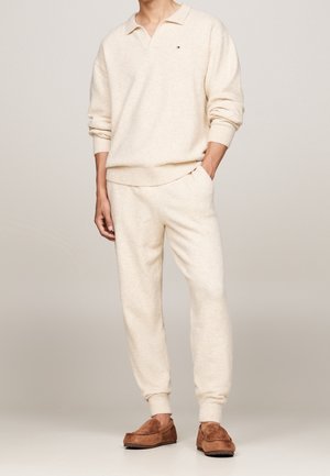 Mann trägt cremefarbenen Strickpullover mit Kragen, passende Jogginghose und braune Wildleder-Mokassinschuhe, steht mit einer Hand in der Tasche.