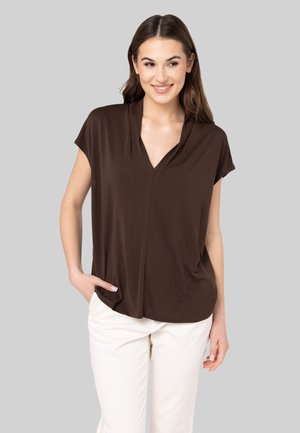 Blouse courte marron à manches courtes avec un col en V et un tissu doux, présentant une coupe décontractée et un drapé subtil, associée à un pantalon de couleur claire.