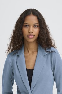 Tânără cu păr lung și creț, de culoare închisă, purtând un sacou albastru deschis peste un top negru, uitându-se direct în cameră pe un fundal simplu.