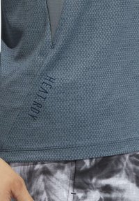 Acercamiento a una camisa azul texturizada con el texto "HEAT.RDY" cerca del dobladillo inferior y pantalones cortos suaves de color gris claro con pequeñas perforaciones.